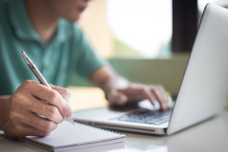 person taking notes with a pen while using a laptop